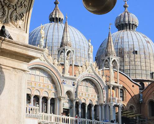 View through an intricately decorated stone archway of the domes and golden crosses of St. Mark's Basilica in Venice under a clear blue sky.