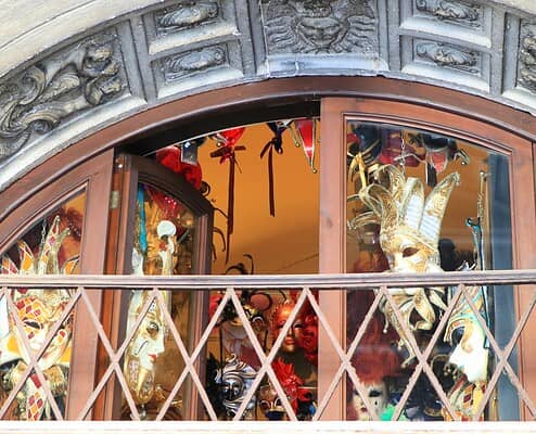 A view through a historic arched window at an artistic selection of Venetian Carnival masks in a shop in Venice.