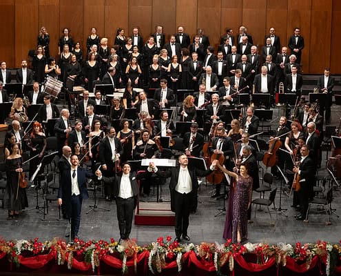 Schlussapplaus beim Neujahrskonzert in Triest • Foto Fabio Parenzan Ein großes Orchester und ein Chor stehen auf einer Bühne mit Holzvertäfelung. Im Vordergrund verbeugen sich vier Solisten händehaltend vor einem festlich geschmückten Bühnenrand.