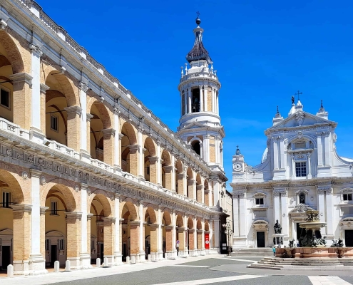 View of the Piazza della Madonna in Loreto, Italy, featuring the white facade of the Basilica della Santa Casa, the Vanvitelli bell tower, and the Apostolic Palace with its grand arched porticos.
