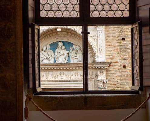 Window view of the San Domenico Portal, Urbino • m24o A view through an open Renaissance window with leaded glass panes looking onto the ornate stone portal of San Domenico church in Urbino, featuring a white terracotta relief.