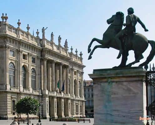 The grand baroque facade of Palazzo Madama in Turin with a bronze equestrian statue in the foreground under a clear blue sky.