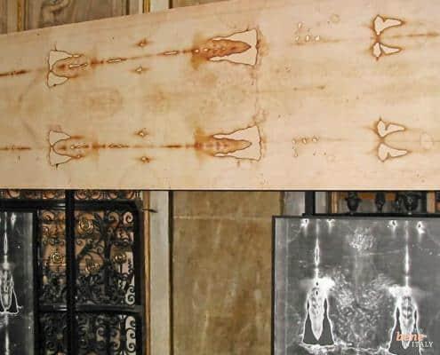 An exhibition of the Shroud of Turin showing the original light-colored linen cloth with burn marks and body imprints above, and two large photographic negative enlargements of the face and torso below.