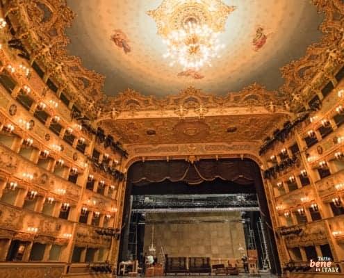 The magnificent, gold-decorated auditorium of Teatro La Fenice in Venice, showing the private boxes, the grand chandelier, and the stage during setup.