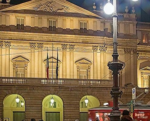 The illuminated Teatro alla Scala in Milan at night with pedestrians in the piazza and a passing red tram in the foreground.