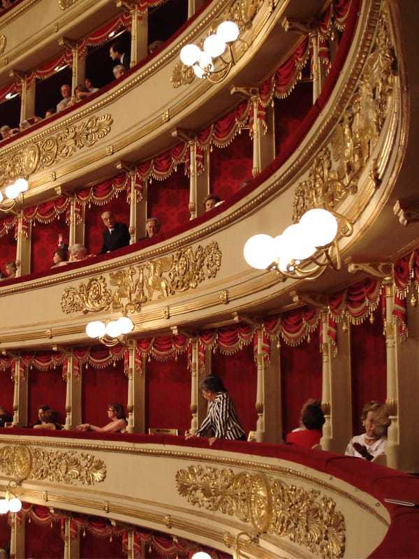 View of the elegant, gold-decorated private boxes of the Teatro alla Scala in Milan featuring red velvet interiors and opera guests during a performance.