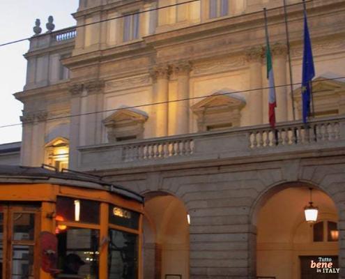 Milanese Charm: Teatro alla Scala and the Historic Tram • m24o The illuminated facade of Teatro alla Scala in Milan at dusk, with a historic yellow tram passing by in the foreground.