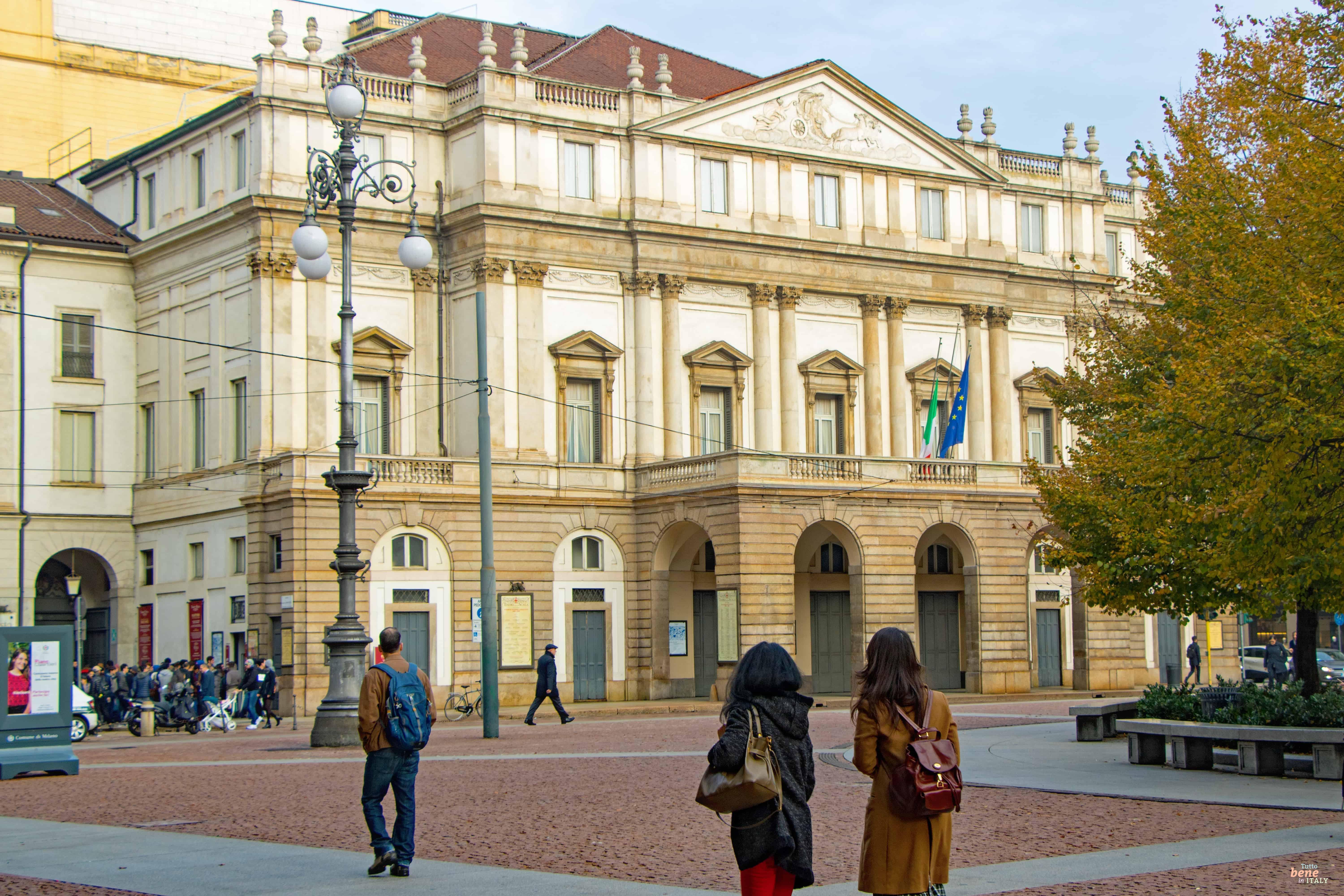 Zwei junge Frauen überqueren die Piazza della Scala in Mailand vor dem Hintergrund des Teatro alla Scala bei klarem Himmel.