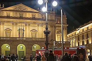 As the lights dim across Piazza della Scala, the magic begins. Join the stylish crowd on their way to the opera house and soak in the vibrant nightly atmosphere of Milan. The illuminated Teatro alla Scala in Milan at night with pedestrians in the piazza and a passing red tram in the foreground.