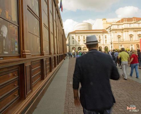 Kunst und Mode auf der Piazza della Scala – Mailänder Flair erleben • m24o Fußgänger gehen an einer künstlerischen Holzinstallation auf der Piazza della Scala in Mailand vorbei; im Hintergrund ist das Opernhaus zu sehen.