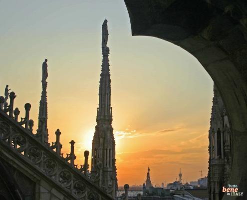 Blick durch einen Steinbogen auf die gotischen Turmspitzen und Statuen des Mailänder Doms während eines goldenen Sonnenuntergangs.