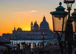 Silhouette of St. Mary of Health Basilica (Santa Maria della Salute) in Venice during a vibrant orange sunset, framed by historic street lamps in the foreground.
