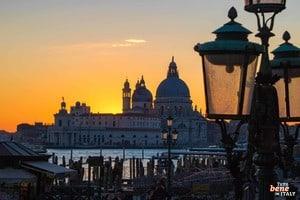 Abendstimmung in Venedig – Sonnenuntergang über der Salute-Kirche • m24o Silhouette der Basilika Santa Maria della Salute in Venedig bei einem leuchtend orangefarbenen Sonnenuntergang, eingerahmt von historischen Straßenlaternen im Vordergrund.