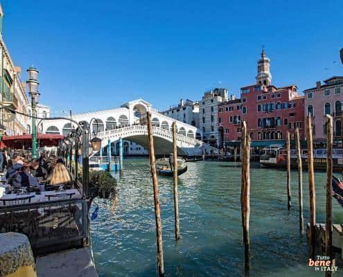 Dining with a View: The Rialto Bridge in Venice • m24o View from a riverside restaurant on the Grand Canal looking towards the Rialto Bridge in Venice under a clear blue sky.