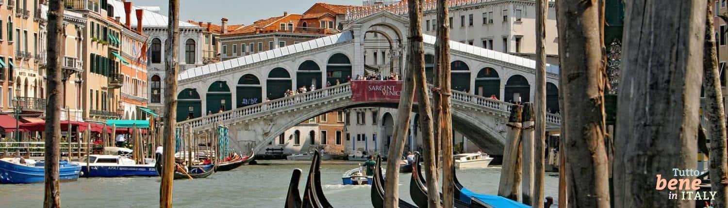 Das Herz von Venedig: Die Rialtobrücke über den Canal Grande Blick durch die hölzernen Gondelstangen auf die historische Rialtobrücke über den Canal Grande in Venedig unter strahlend blauem Himmel.