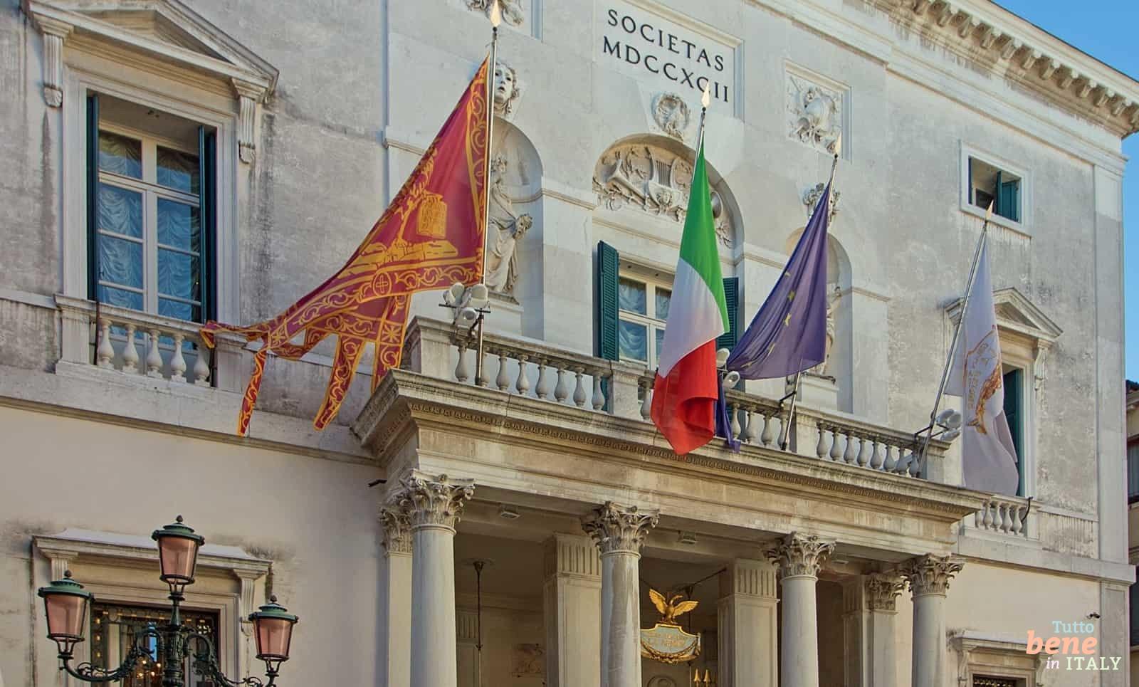 Die prachtvolle Fassade des Teatro La Fenice in Venedig mit wehenden Flaggen und klassizistischen Skulpturen unter blauem Himmel.
