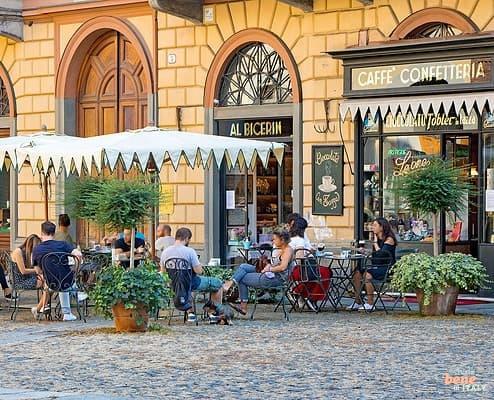 The historic exterior of Caffè Al Bicerin in Turin with customers sitting at small outdoor tables under white umbrellas on a cobblestone square, next to a vintage dark wood storefront.