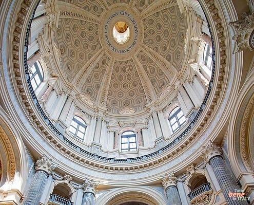 Innenraum und Kuppel der Basilica di Superga in Turin • m24o Prachtvoller barocker Innenraum der Basilica di Superga in Turin mit Blick nach oben in die monumentale, hellbeige Kuppel mit Wabenmuster, flankiert von hohen, kannelierten Säulen aus grauem Marmor und Besuchern im Kirchenschiff.