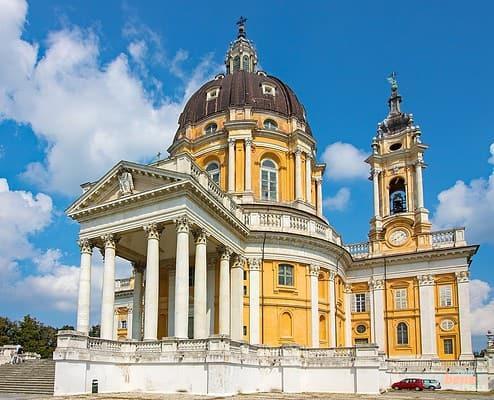 Außenansicht der Basilica di Superga in Turin bei Sonnenschein • m24o Die barocke Basilica di Superga in Turin mit ihrer monumentalen dunkelbraunen Kuppel, der ockergelben Fassade, einer weißen Säulenvorhalle und einem eleganten Glockenturm unter strahlend blauem Himmel mit weißen Wolken.
