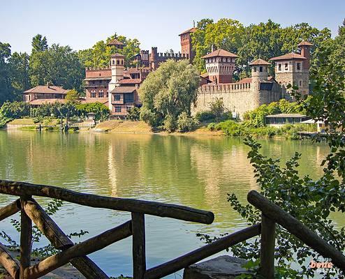 Turin Valentino Park mit dem mittelalterlichen Dorf • m24o Das mittelalterliche Dorf Borgo Medievale in Turin mit seinen Festungsmauern und Türmen aus Backstein, eingebettet in grüne Bäume am Ufer des Flusses Po, der die Szenerie spiegelt, fotografiert hinter einem Holzgeländer im Vordergrund.
