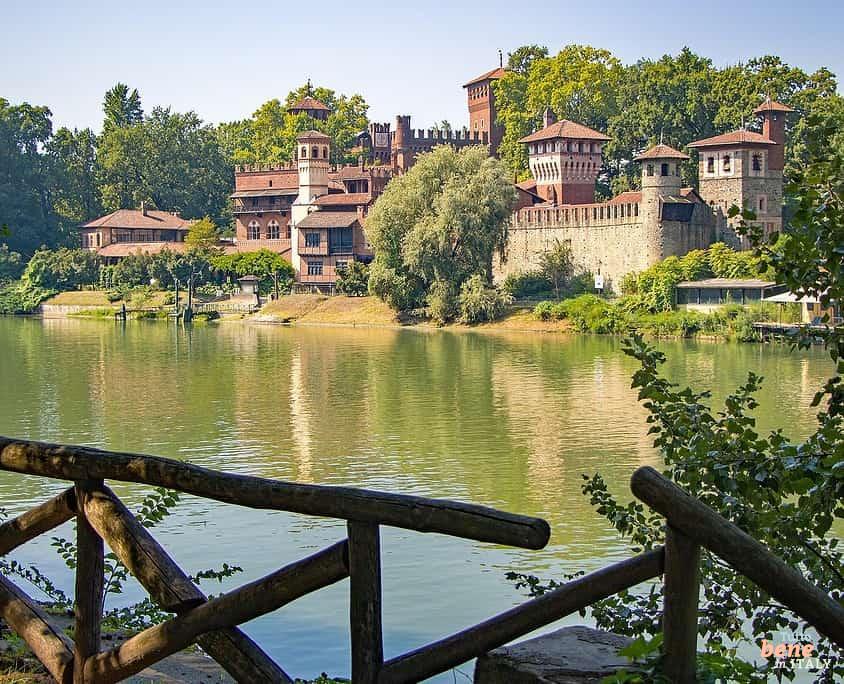 Turin Valentino Park mit dem mittelalterlichen Dorf • m24o Das mittelalterliche Dorf Borgo Medievale in Turin mit seinen Festungsmauern und Türmen aus Backstein, eingebettet in grüne Bäume am Ufer des Flusses Po, der die Szenerie spiegelt, fotografiert hinter einem Holzgeländer im Vordergrund.