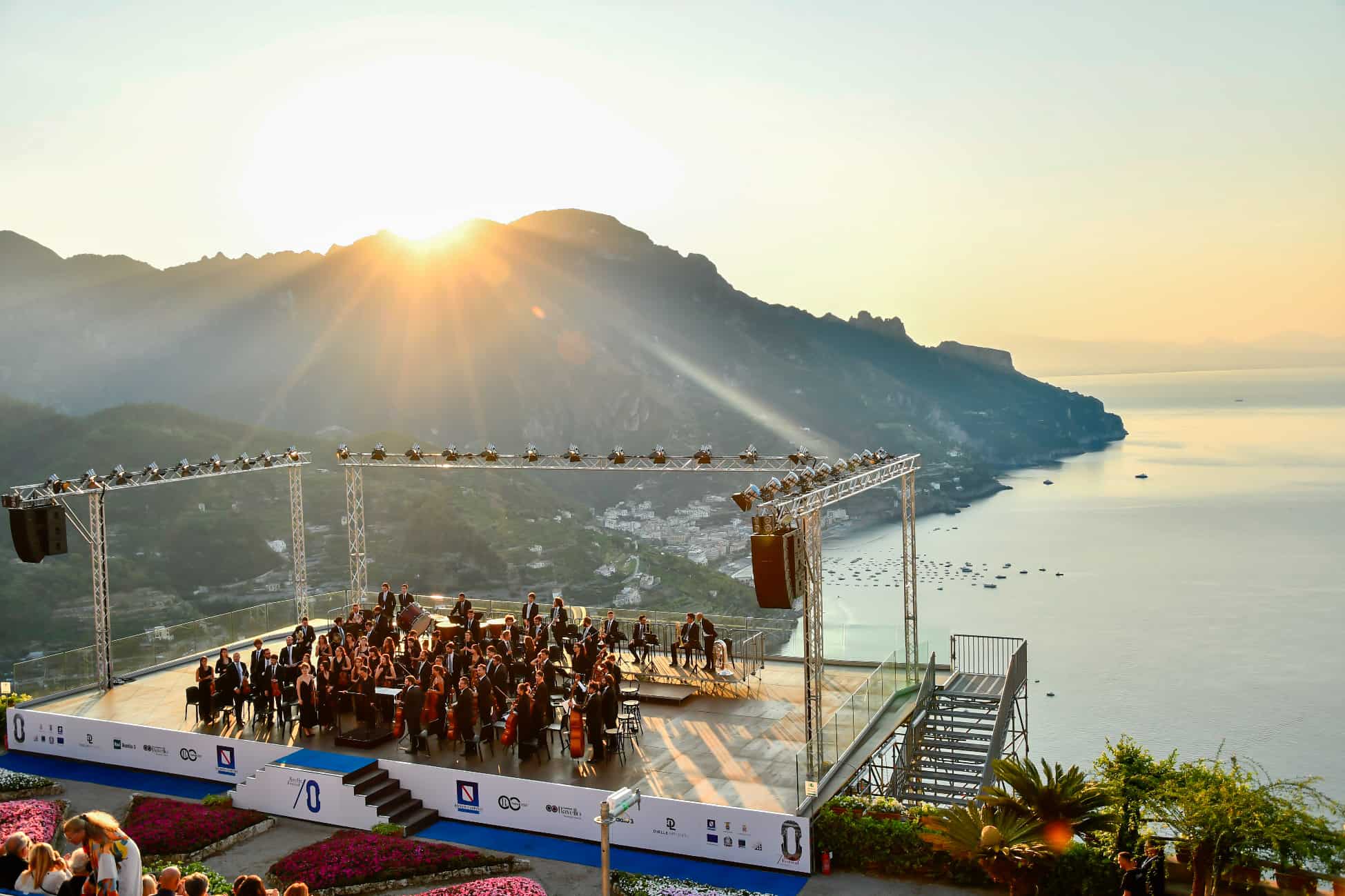 A symphony orchestra performs at the Ravello Festival on the Villa Rufolo Belvedere stage as the sun rises brightly over the Amalfi Coast mountains.