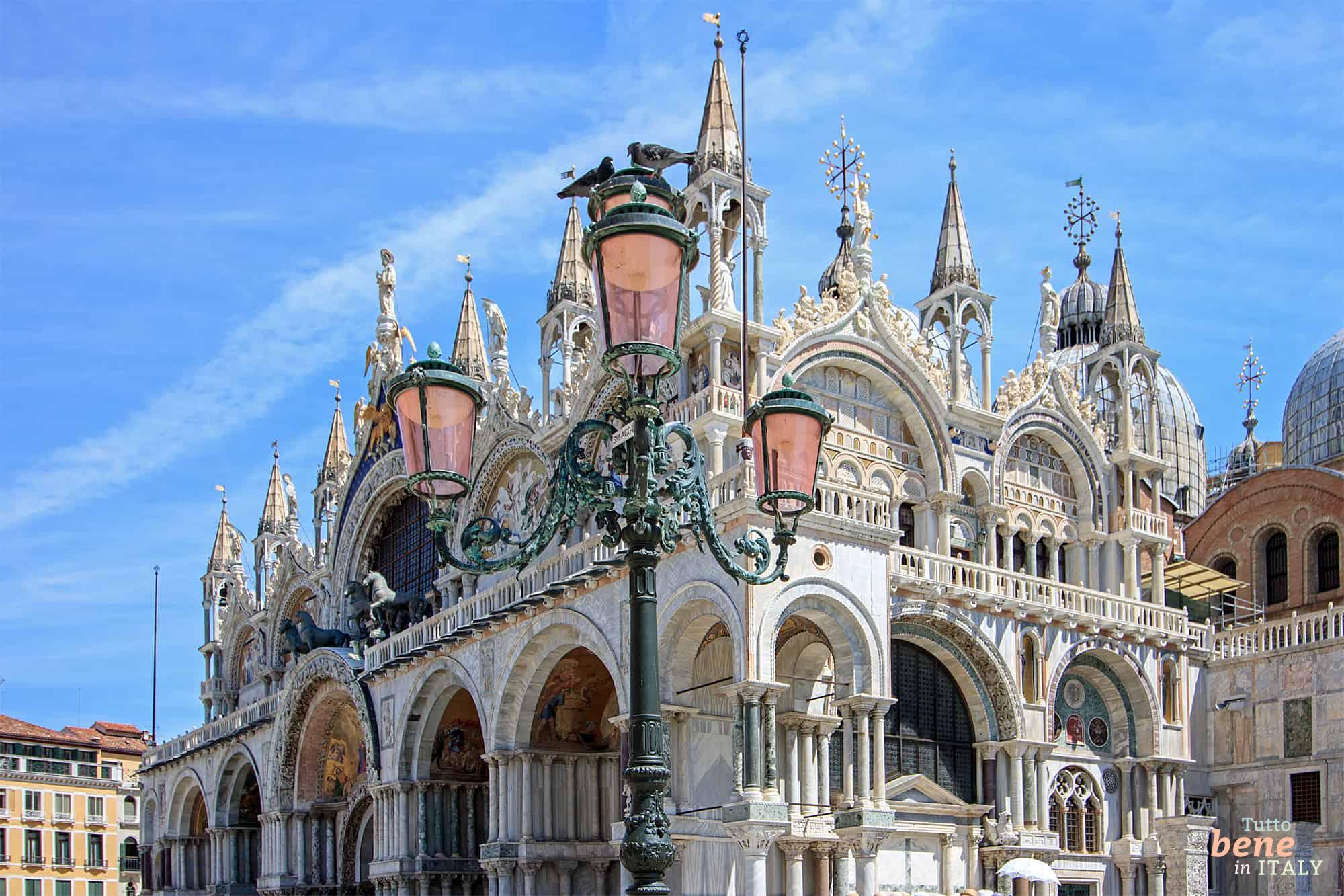 Die prunkvolle Fassade des Markusdoms (Basilica di San Marco) in Venedig mit seinen goldenen Mosaiken und einer historischen Stra&szlig;enlaterne im Vordergrund.