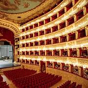 Innenansicht des historischen Opernhauses Teatro di San Carlo in Neapel • m24o Ein prunkvoller, hufeisenförmiger Opernsaal mit sechs Rängen goldverzierter Logen und roten Samtsitzen, die auf eine beleuchtete Bühne blicken. An der Decke ist ein großes Fresko zu sehen.
