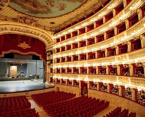 Innenansicht des historischen Opernhauses Teatro di San Carlo in Neapel • m24o Ein prunkvoller, hufeisenförmiger Opernsaal mit sechs Rängen goldverzierter Logen und roten Samtsitzen, die auf eine beleuchtete Bühne blicken. An der Decke ist ein großes Fresko zu sehen.