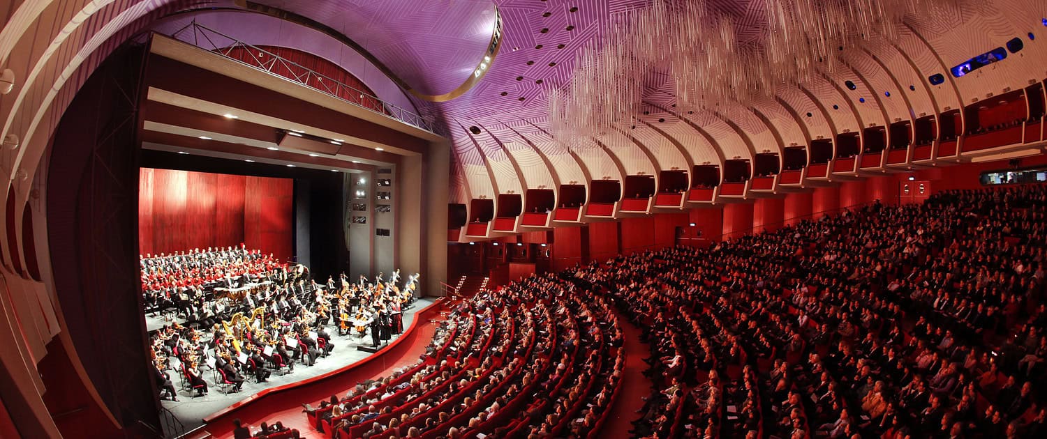 Blick auf die Bühne mit Orchester und Chor im voll besetzten Zuschauerraum des Teatro Regio di Torino mit moderner lila Decke.