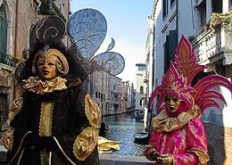 Zwei prächtig maskierte Gestalten in aufwendigen schwarzen und pinkfarbenen Barock-Kostümen auf einer Brücke über einem Kanal in Venedig.