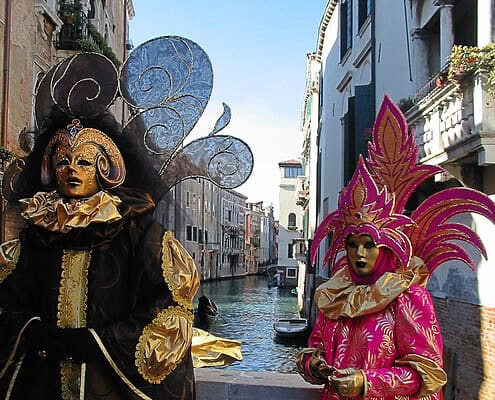 Two magnificently masked figures in elaborate black and pink Baroque costumes on a bridge overlooking a canal in Venice.