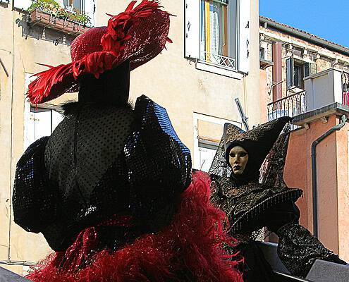 Elegance and Mystery: Mask Magic at the Venice Carnival • m24o Two masked figures at the Venice Carnival: A person in an elaborate black sequined costume with a red feathered hat in the foreground and another mask in gold and black in the background.