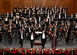 Ein großes Orchester und ein Chor stehen auf einer Bühne mit Holzvertäfelung. Im Vordergrund verbeugen sich vier Solisten händehaltend vor einem festlich geschmückten Bühnenrand.