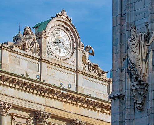 Nahaufnahme der Fassade des Palazzo della Veneranda Fabbrica del Duomo in Mailand mit einer großen monumentalen Uhr und Statuen, daneben die gotische Fassade des Mailänder Doms mit Heiligenfiguren.