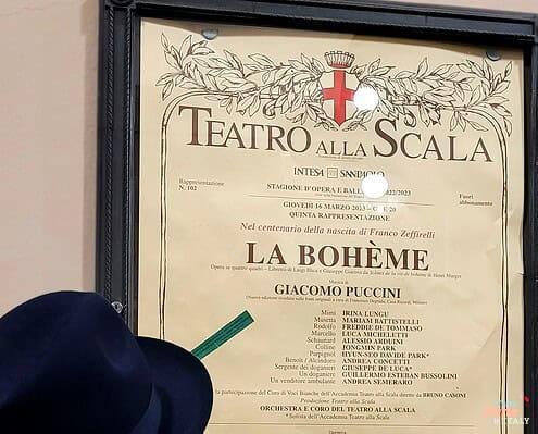 A man in a hat studying the official opera poster for La Bohème by Giacomo Puccini at Teatro alla Scala.