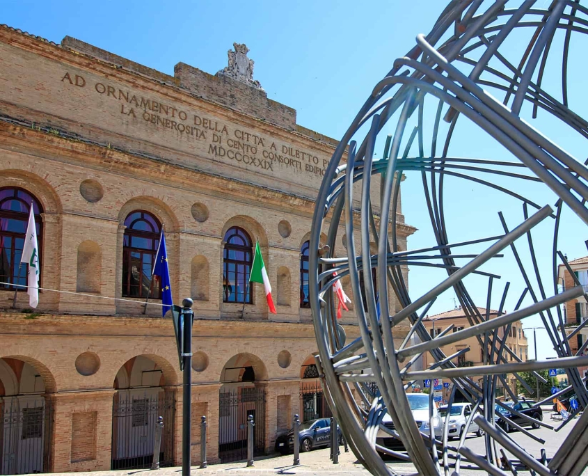 Entrance of the Sferisterio in Macerata with Sculpture • m24o The monumental neoclassical facade of the Sferisterio in Macerata featuring a sculpture in the foreground, promoting the Opera Festival.