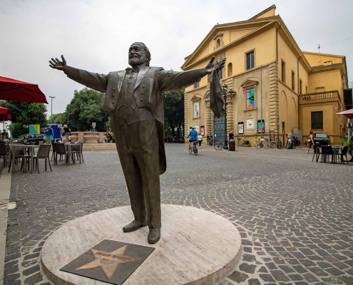 Bronze statue of tenor Luciano Pavarotti with open arms in a public square in Pesaro, with the Teatro Rossini in the background.