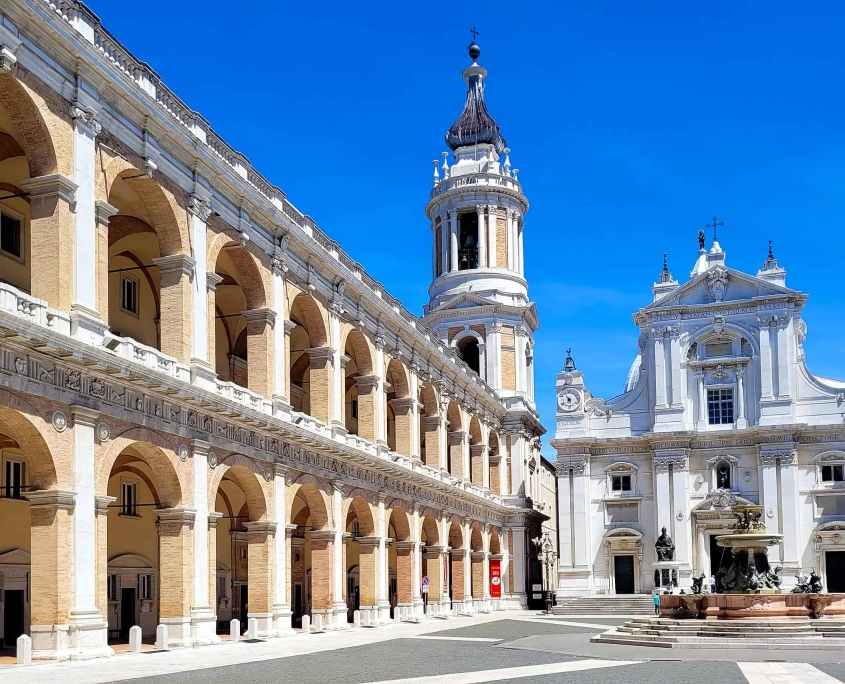 View of the Piazza della Madonna in Loreto, Italy, featuring the white facade of the Basilica della Santa Casa, the Vanvitelli bell tower, and the Apostolic Palace with its grand arched porticos.