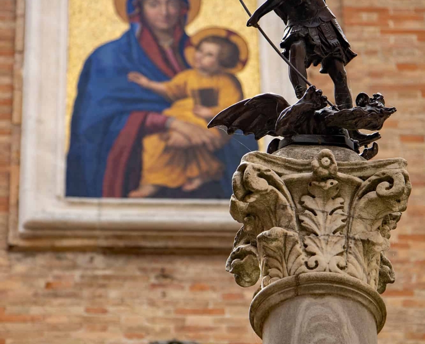 Sacred Detail in Urbino St. George and Madonna Mosaic • m24o Bronze statue of Saint George on a Corinthian column in Urbino, Italy, with a golden Madonna and Child mosaic visible on a brick wall in the background.