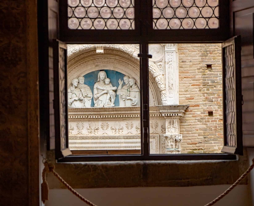 Window view of the San Domenico Portal, Urbino • m24o A view through an open Renaissance window with leaded glass panes looking onto the ornate stone portal of San Domenico church in Urbino, featuring a white terracotta relief.