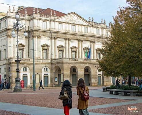 Ein Spaziergang zur Scala – Entdecken Sie Mailand mit m24o • m24o Zwei junge Frauen spazieren über die Piazza della Scala in Mailand mit dem Teatro alla Scala im Hintergrund unter blauem Himmel.