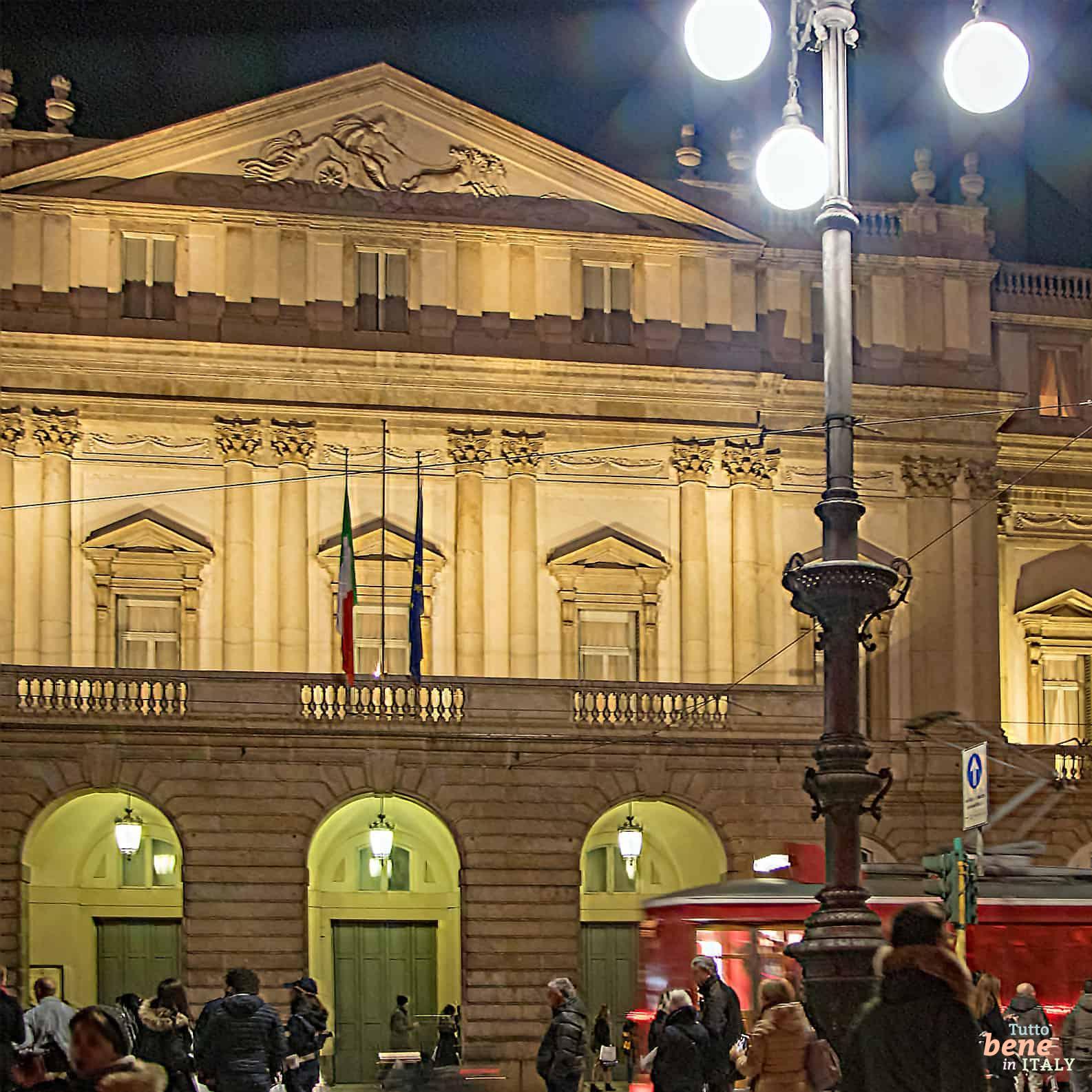 Evening Anticipation: La Scala Awaits Its Audience The illuminated Teatro alla Scala in Milan at night with pedestrians in the piazza and a passing red tram in the foreground.